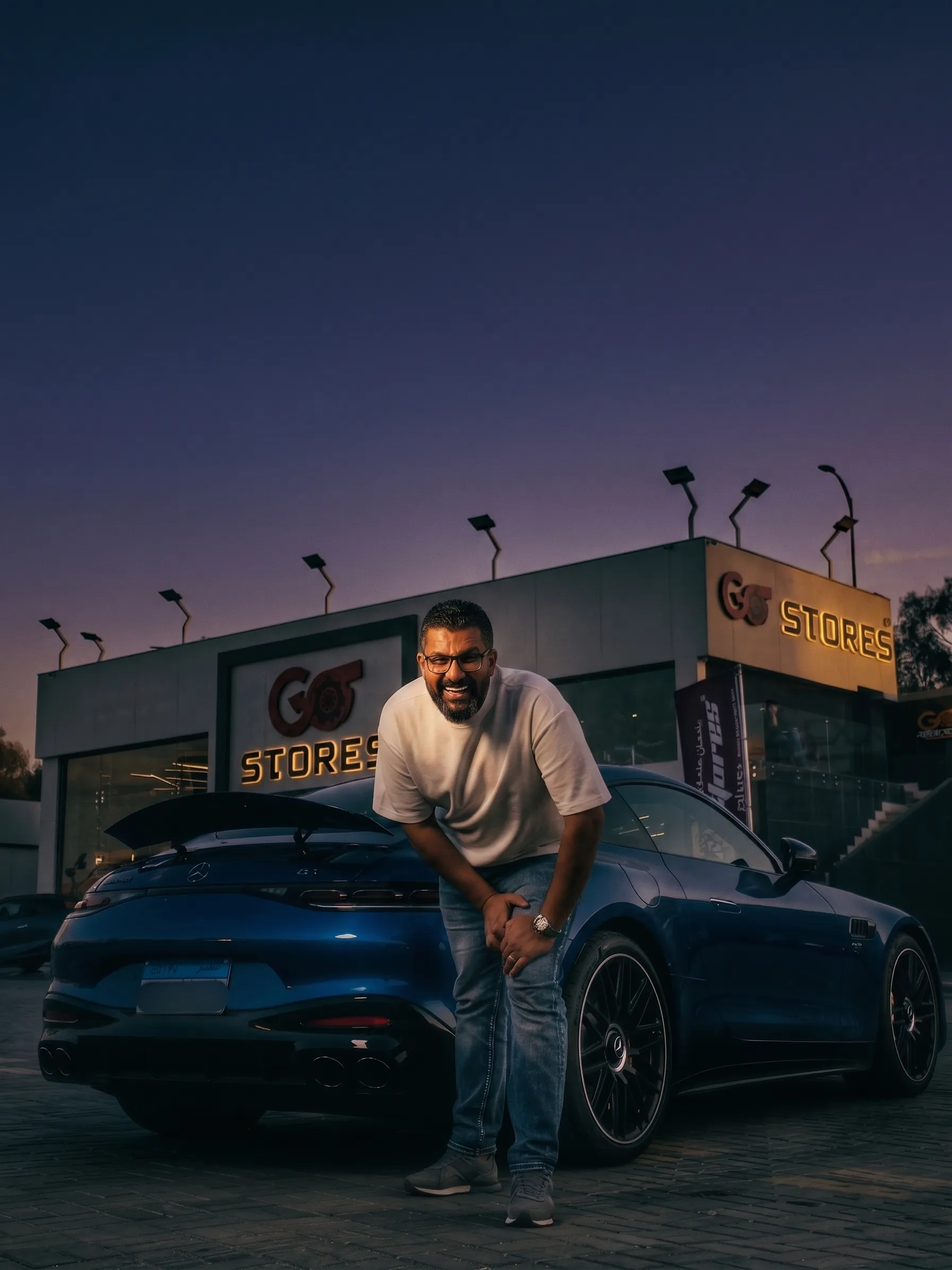 Mohammed Osman, founder of Go Stores, leaning on his blue Porsche in front of the storefront at dusk